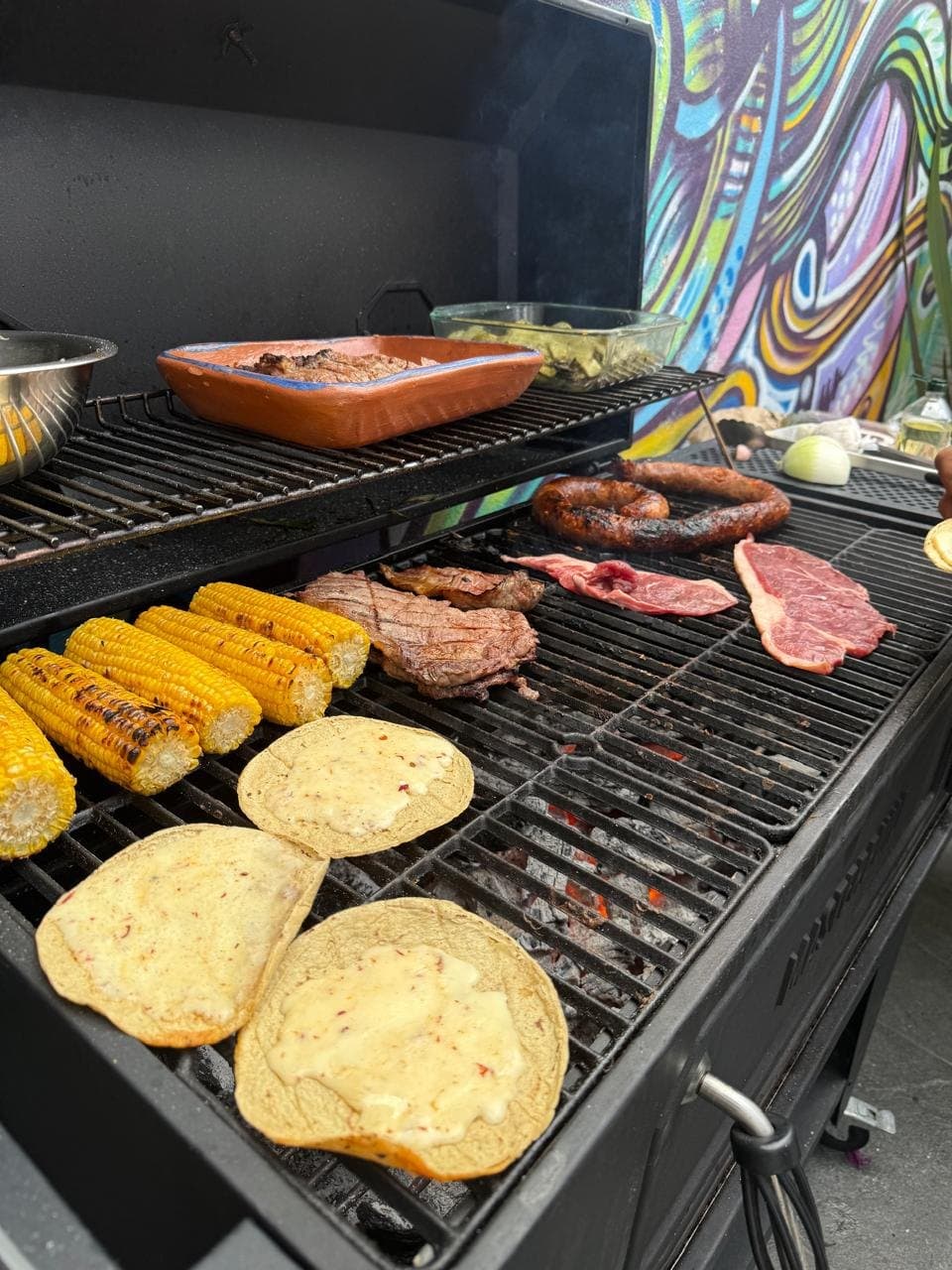 Parrilla de carbón con arrachera, chorizo, elotes y tortillas con queso; se ven las brasas encendidas bajo la rejilla.