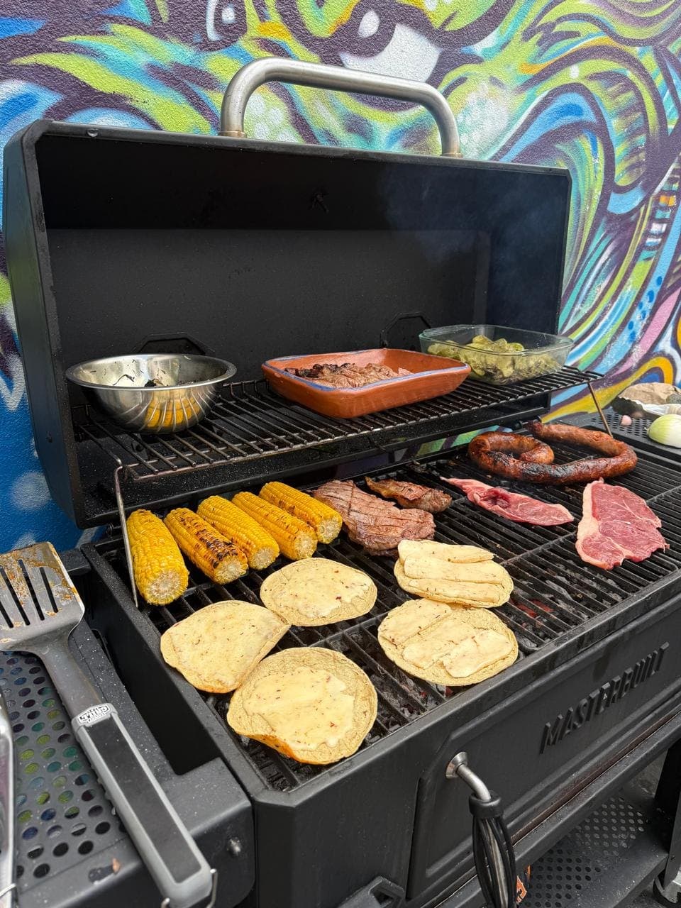 Cortes, chorizo, elotes y tortillas sobre la parrilla, con la tapa abierta y el carbón encendido.