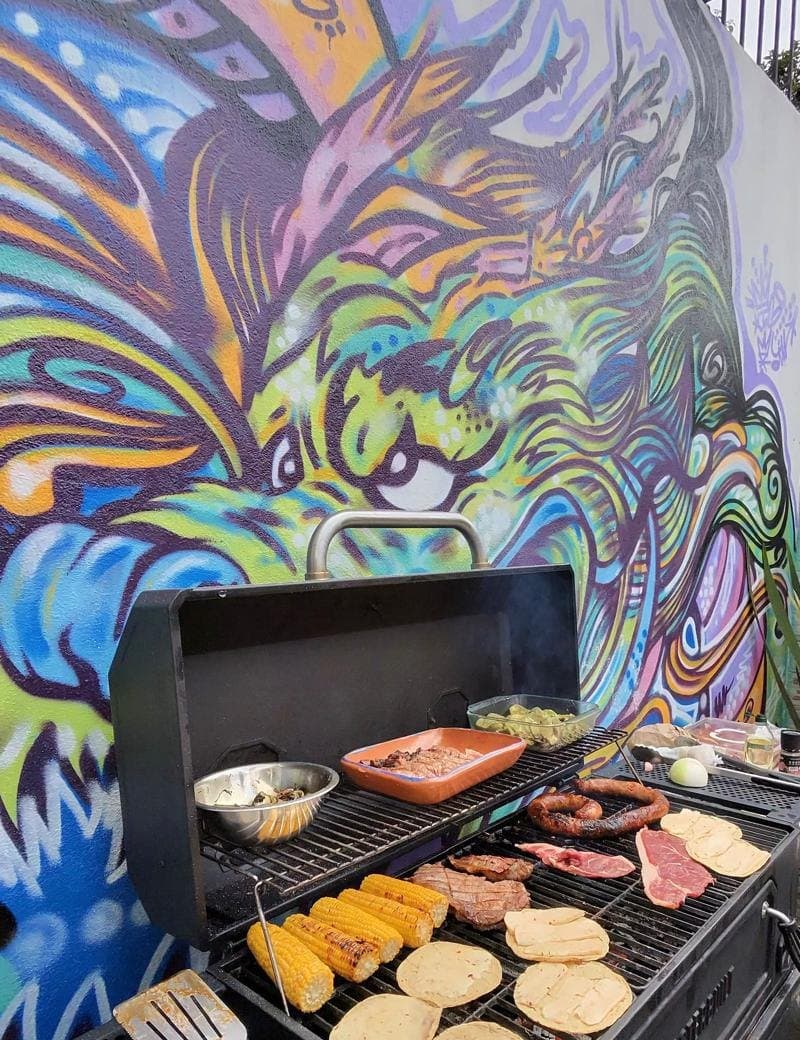 La parrilla encendida en un patio con un mural de colores, con cortes y elotes al fuego.